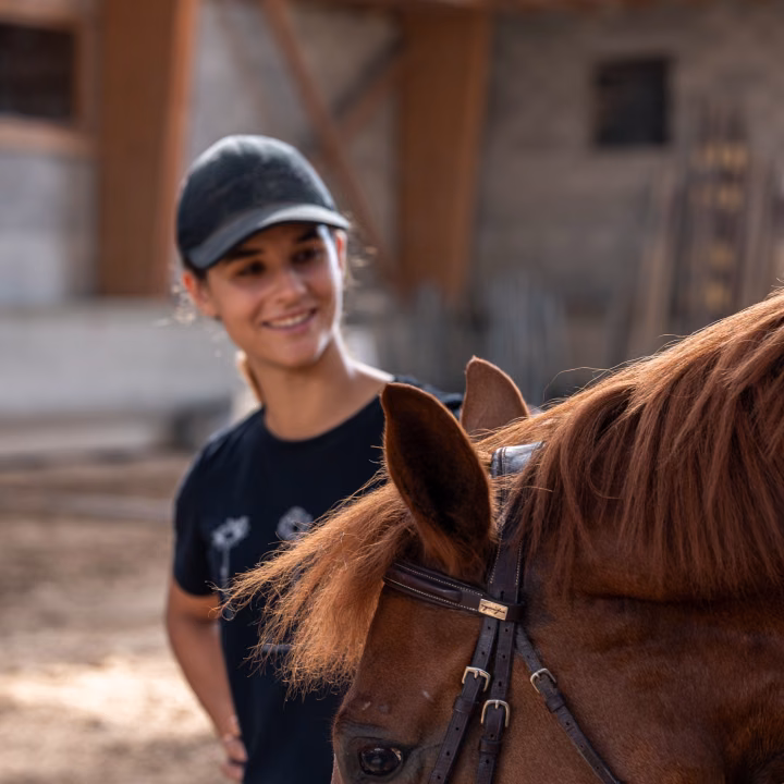 Une monitrice en casquette sourit près de la tête d'un cheval alezan bridé, dans le manège, lumière chaude sur le sable.