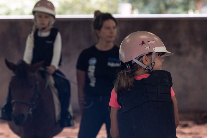 Une fillette casquée en gilet de protection, sur son poney, regarde sur le côté, une monitrice floue derrière elle.