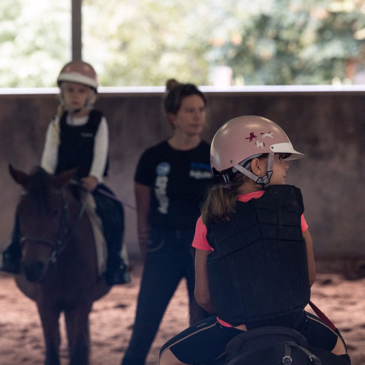 Une fillette casquée en gilet de protection, sur son poney, regarde sur le côté, une monitrice floue derrière elle.