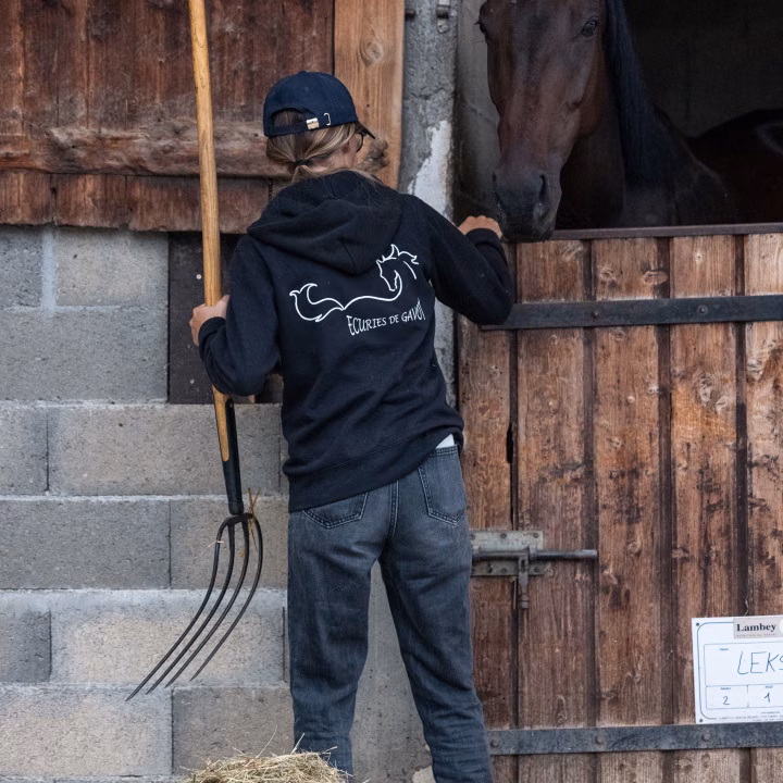 Une personne de dos, en sweat marine à logo, tient une fourche et caresse un cheval bai à la porte de son box.