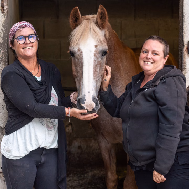 Deux femmes souriantes posent avec un cheval alezan à liste blanche, qu'elles caressent, à la porte de son box.