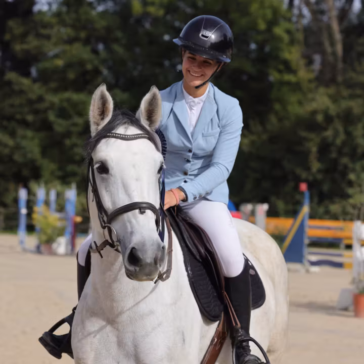 Cavalière souriante en veste bleu ciel sur un poney gris, sur la piste de concours